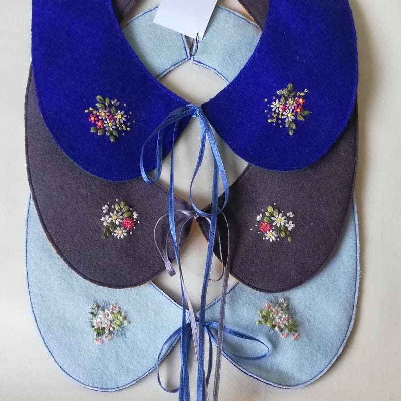 K&ouml;nnte beinhalten: Drei Filzkragen in Blau-, Grau- und Hellblaut&ouml;nen, jeweils mit gestickten Blumenmotiven verziert. Die Kragen sind mit blauen B&auml;ndern zum Binden verbunden. Oben ist ein Etikett sichtbar.