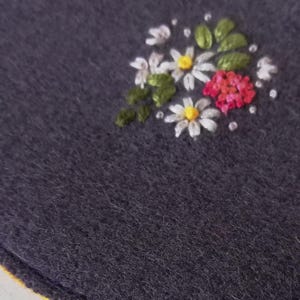 K&ouml;nnte beinhalten: Nahaufnahme eines dunkelgrauen Stoffes mit einem gestickten Blumendesign. Die Stickerei zeigt wei&szlig;e G&auml;nsebl&uuml;mchen mit gelben Zentren, rote Bl&uuml;ten, gr&uuml;ne Bl&auml;tter und kleine wei&szlig;e Punkte. Der Stoff hat ein strukturiertes Aussehen.