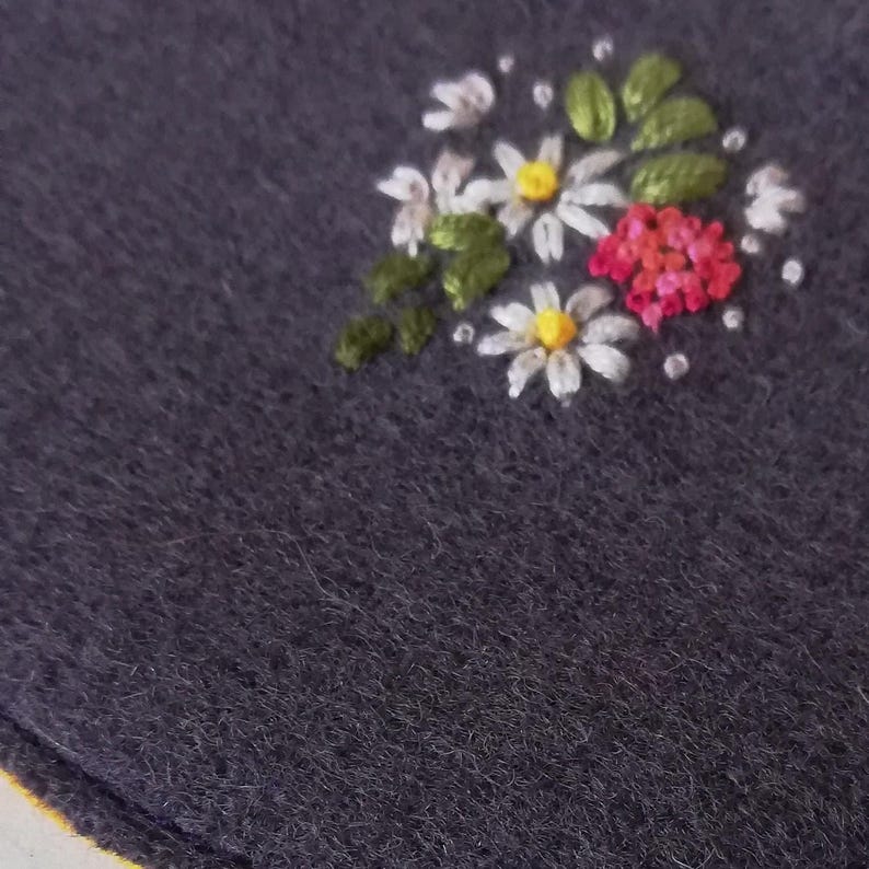 K&ouml;nnte beinhalten: Nahaufnahme eines dunkelgrauen Stoffes mit einem gestickten Blumendesign. Die Stickerei zeigt wei&szlig;e G&auml;nsebl&uuml;mchen mit gelben Zentren, rote Bl&uuml;ten, gr&uuml;ne Bl&auml;tter und kleine wei&szlig;e Punkte. Der Stoff hat ein strukturiertes Aussehen.