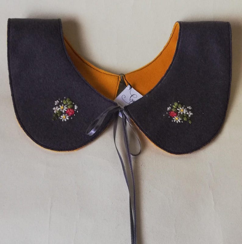 K&ouml;nnte beinhalten: Ein dunkelgrauer Filzkragen mit orangefarbenem Futter und gestickten floralen Details. Der Kragen ist mit einem grauen Band gebunden. Der Kragen hat eine abgerundete Form und ist zum Tragen um den Hals konzipiert.