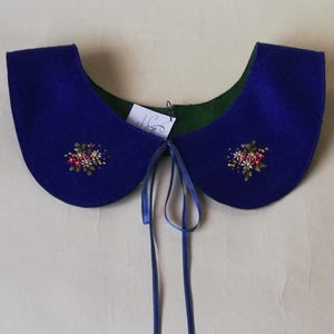K&ouml;nnte beinhalten: Ein k&ouml;nigsblauer Filzkragen mit gr&uuml;ner Unterseite und gestickten floralen Details. Der Kragen hat eine abgerundete Form und wird mit blauen B&auml;ndern gebunden. Ein kleines Etikett ist angebracht.