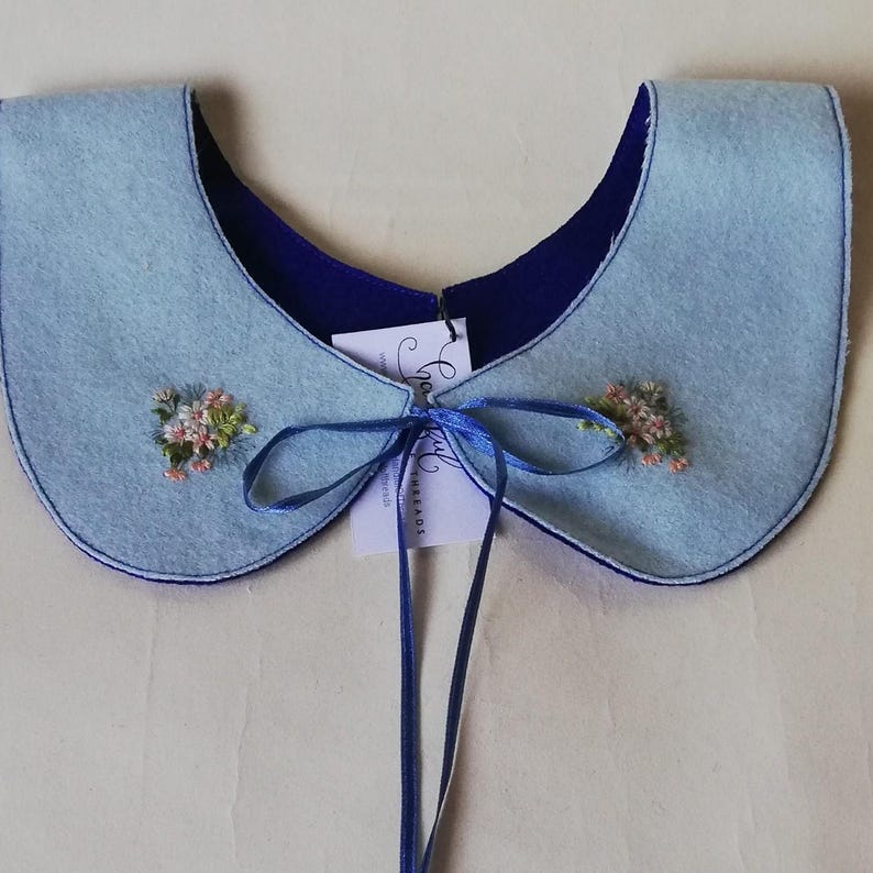 K&ouml;nnte beinhalten: Ein hellblauer Filzkragen mit dunkelblauem Futter und passendem blauem Band. Der Kragen hat kleine gestickte Blumenakzente in Rosa und Wei&szlig;. Ein Etikett ist angebracht.
