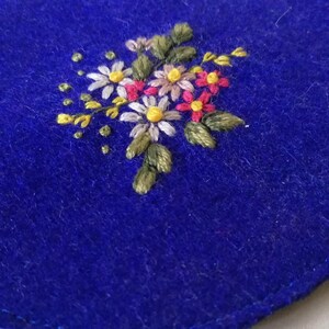 K&ouml;nnte beinhalten: Nahaufnahme eines leuchtend blauen Stoffes mit einem gestickten Blumendesign. Die Stickerei zeigt einen Strau&szlig; kleiner Blumen in Wei&szlig;-, Rosa-, Gelb- und Braunt&ouml;nen mit gr&uuml;nen Bl&auml;ttern und Stielen. Die Textur des Stoffes ist sichtbar.