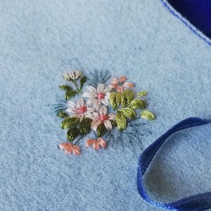 K&ouml;nnte beinhalten: Nahaufnahme eines hellblauen Stoffes mit einem gestickten Blumenmuster. Die Stickerei zeigt kleine Bl&uuml;ten in Rosa, Wei&szlig; und Pfirsich, mit gr&uuml;nen Bl&auml;ttern. Ein blaues Band ist ebenfalls zu sehen, was auf ein Bastelprojekt oder ein dekoratives Element hindeutet.