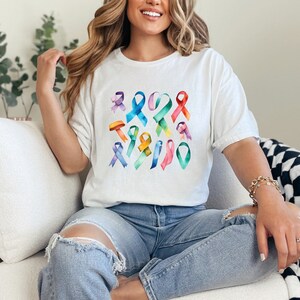 Peut inclure: Un t-shirt blanc avec un motif color&eacute; de diff&eacute;rentes formes de rubans. Les rubans sont dans des tons de bleu, vert, violet, rose, jaune et orange. Le t-shirt est port&eacute; par une personne aux longs cheveux bruns.