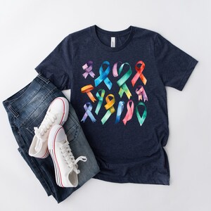 Peut inclure: Un t-shirt bleu marine avec un motif aquarelle color&eacute; de formes de rubans. Le t-shirt est pr&eacute;sent&eacute; sur un fond blanc avec une paire de baskets blanches et un jean bleu.