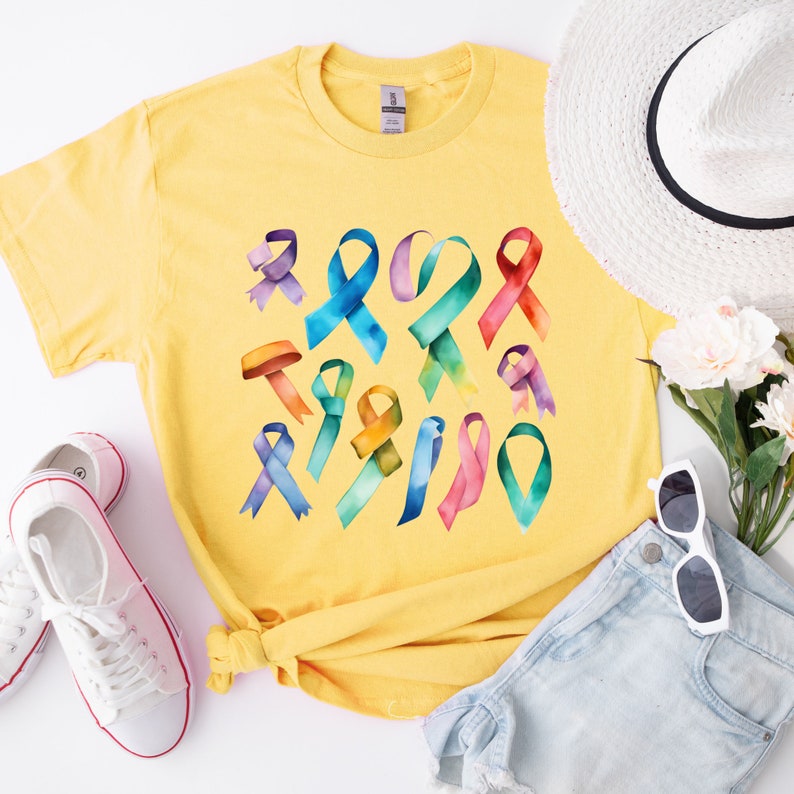 Peut inclure: Un t-shirt jaune avec un motif de plusieurs rubans aquarelle dans diff&eacute;rentes couleurs. Les rubans sont dispos&eacute;s de mani&egrave;re al&eacute;atoire et sont tous nou&eacute;s en un n&oelig;ud.