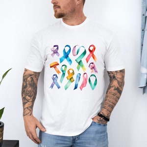 Peut inclure: Un t-shirt blanc avec un motif aquarelle color&eacute; de plusieurs rubans de sensibilisation. Les rubans sont dans diff&eacute;rentes nuances de bleu, vert, jaune, orange, rose et violet.