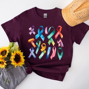 Peut inclure: Un t-shirt marron avec un motif aquarelle color&eacute; de plusieurs formes de rubans. Les rubans sont dans des tons de rose, bleu, vert, jaune et violet.