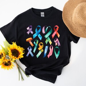 Peut inclure: T-shirt noir avec un motif aquarelle color&eacute; de plusieurs rubans de sensibilisation. Les rubans sont dans des tons de rose, bleu, vert, jaune et violet.