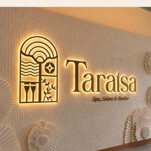 May include: A wall-mounted sign featuring the text Taraisa Spa, Salon & Studio in gold lettering with backlighting. To the left is a rectangular panel with an arched top containing geometric line art. The wall has a textured finish.