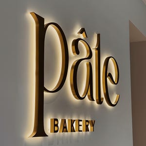May include: A wall-mounted sign for a bakery, featuring the word "Pâte" in elegant gold lettering with a warm, glowing backlight. Below, the word "BAKERY" is displayed in a similar gold font. The sign is mounted on a light gray wall.