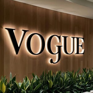 May include: A black and white sign with the word "VOGUE" in large, bold letters. The sign is illuminated with white lights and is mounted on a wood panel wall.