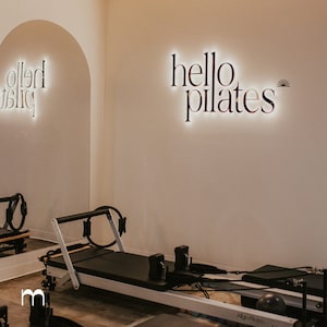 May include: A white wall with a lit sign that says "hello pilates" in black lettering. A pilates reformer is in the foreground with a black mat and a black exercise ball.