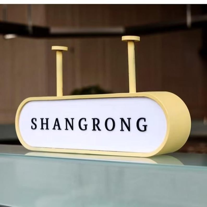 Op de afbeelding: Een geel en wit bord met het woord "SHANGRONG" in zwarte letters. Het bord heeft afgeronde randen en wordt opgehangen aan twee gele palen.