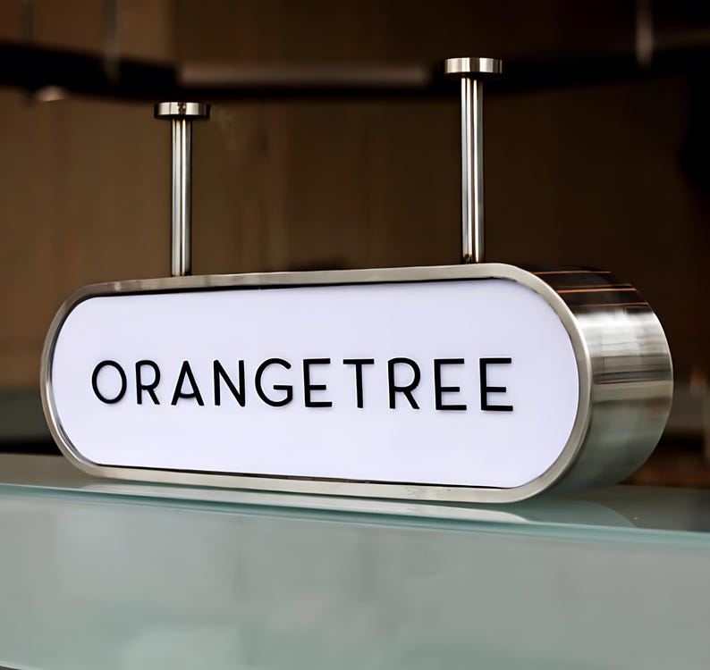 Op de afbeelding: Een rechthoekig bord met afgeronde randen, met het woord "ORANGETREE" in zwarte hoofdletters op een witte achtergrond. Het bord is omlijst door geborsteld metaal en hangt aan het plafond met twee metalen staven.