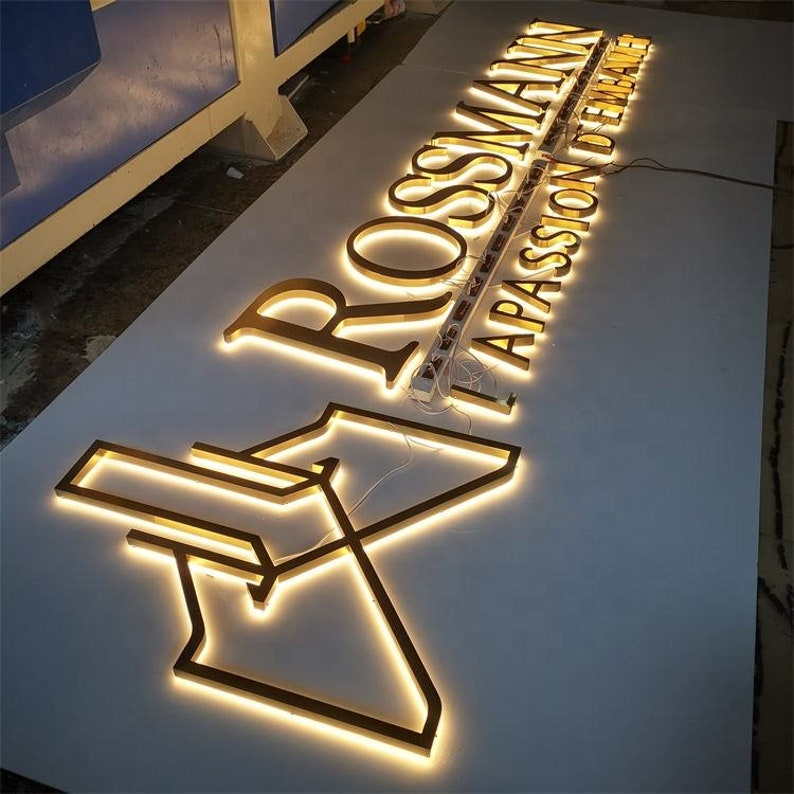 May include: Gold-colored metal sign with the words "ROSSMANN" and "APASSION FOR HOME" illuminated with white LED lights.
