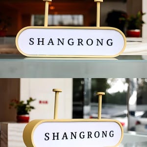Op de afbeelding: Een gele rechthoekige bewegwijzering met het woord "SHANGRONG" in zwarte hoofdletters. De bewegwijzering heeft een witte achtergrond en wordt opgehangen door twee verticale palen. De bewegwijzering wordt binnenshuis weergegeven.