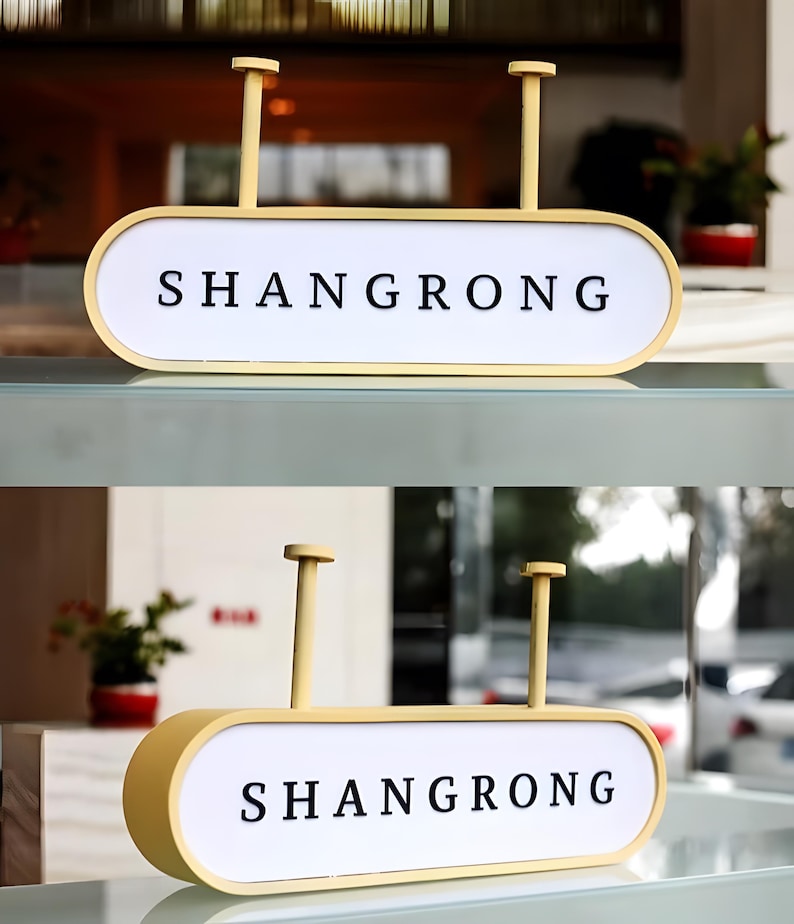 Op de afbeelding: Een gele rechthoekige bewegwijzering met het woord "SHANGRONG" in zwarte hoofdletters. De bewegwijzering heeft een witte achtergrond en wordt opgehangen door twee verticale palen. De bewegwijzering wordt binnenshuis weergegeven.