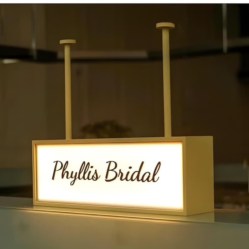 Op de afbeelding: Een rechthoekig, verlicht bord met de tekst "Phyllis Bridal" in elegant schrift. Het bord is cr&egrave;mekleurig en wordt door twee verticale staven aan het plafond gehangen. De achtergrond is zacht verlicht, wat een warme sfeer cre&euml;ert.