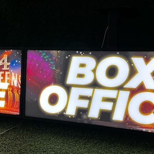 Op de afbeelding: Twee verlichte borden met de tekst "BOX OFFICE" in grote witte letters. De borden tonen ook "4 QUEENS" en kleurrijke graphics, waarschijnlijk voor een show. De borden zijn op een donkere achtergrond gemonteerd.