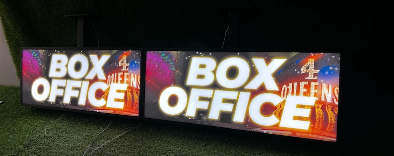 Op de afbeelding: Twee verlichte borden met de tekst "BOX OFFICE" in grote witte letters. De borden tonen ook "4 QUEENS" en kleurrijke graphics, waarschijnlijk voor een show. De borden zijn op een donkere achtergrond gemonteerd.