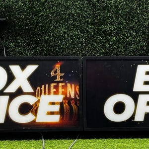 Op de afbeelding: Twee rechthoekige zwarte kassa-borden met witte verlichte tekst "BOX OFFICE". De borden hebben een donkere achtergrond met een kleurrijke afbeelding en de tekst "4 QUEENS". De borden zijn gemonteerd op een groene ondergrond.