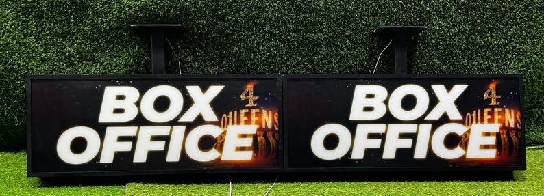 Op de afbeelding: Twee rechthoekige zwarte kassa-borden met witte verlichte tekst "BOX OFFICE". De borden hebben een donkere achtergrond met een kleurrijke afbeelding en de tekst "4 QUEENS". De borden zijn gemonteerd op een groene ondergrond.