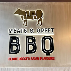 May include: A sign with a gold silhouette of a cow and the text "Meats & Greet BBQ Flame-Kissed Asian Flavours".