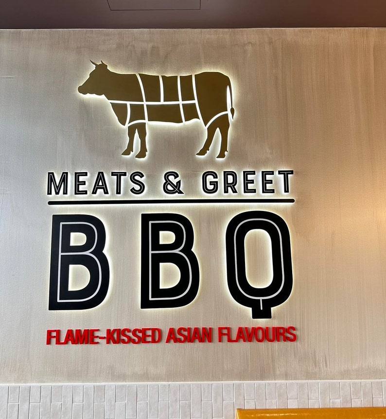 May include: A sign with a gold silhouette of a cow and the text "Meats & Greet BBQ Flame-Kissed Asian Flavours".
