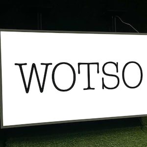 Op de afbeelding: Een rechthoekig wit bord met het woord "WOTSO" in vette, zwarte letters. Het bord is omlijst in een donkere kleur en staat tegen een donkere achtergrond met groen gras.