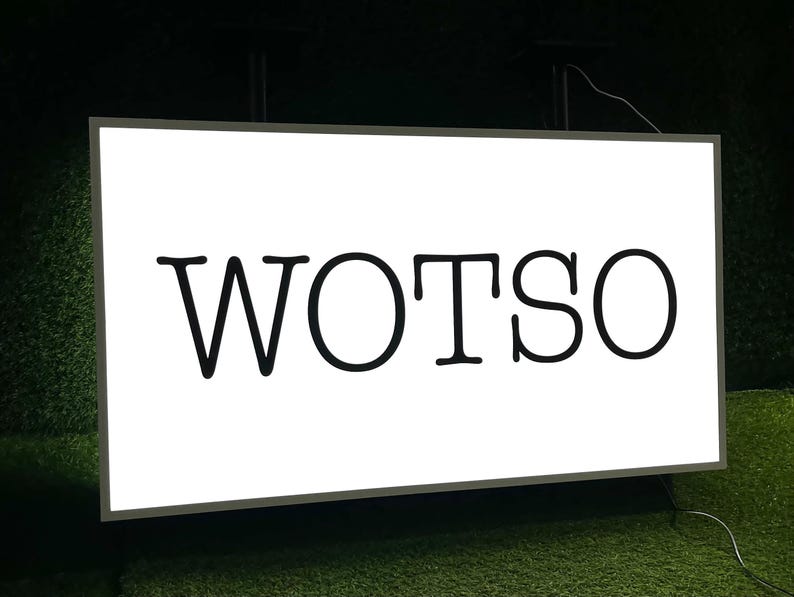 Op de afbeelding: Een rechthoekig wit bord met het woord "WOTSO" in vette, zwarte letters. Het bord is omlijst in een donkere kleur en staat tegen een donkere achtergrond met groen gras.
