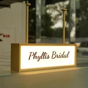 Op de afbeelding: Een rechthoekig, verlicht bord met de tekst "Phyllis Bridal" in een cursief lettertype. Het bord is ingekaderd in een lichtgekleurde lijst en wordt opgehangen door twee verticale palen. De achtergrond is wazig, wat wijst op een binnenomgeving.