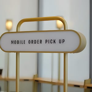 Op de afbeelding: Een goudkleurig bord met de tekst "MOBILE ORDER PICK UP" in zwarte letters. Het bord is verlicht en gemonteerd op een goudkleurig metalen frame. Het bord bevindt zich in een moderne restaurantomgeving.