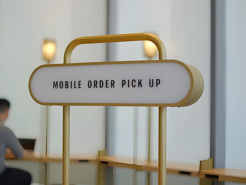 Op de afbeelding: Een goudkleurig bord met de tekst "MOBILE ORDER PICK UP" in zwarte letters. Het bord is verlicht en gemonteerd op een goudkleurig metalen frame. Het bord bevindt zich in een moderne restaurantomgeving.