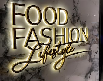 May include: Gold-colored sign with the words "FOOD FASHION LIFESTYLE" in a modern font. The sign is illuminated with white lights.