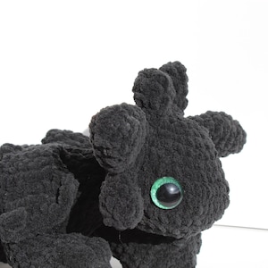 May include: A black crocheted plush toy dragon with green eyes. The dragon is lying on its side with its head turned to the right.