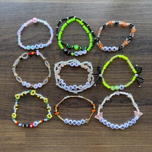 Peut inclure: Neuf bracelets en perles colorées avec différents mots et motifs. Les bracelets sont faits de petites perles colorées et présentent des mots comme "Lando", "LHA", "FC43" et "Lando Wins".