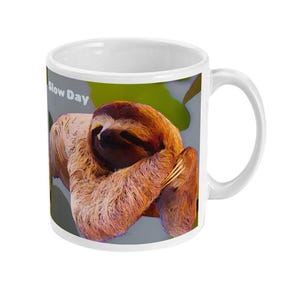 May include: White ceramic mug with a cartoon sloth illustration. The sloth has brown fur and is resting on a branch. The mug has the text "Slow Day". The background is green and grey.