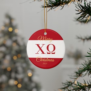 Può includere: Ornamento natalizio rotondo con un design rosso e bianco. L'ornamento presenta le parole "Merry Christmas" e le lettere greche "XΩ" in oro. È incluso anche l'anno "2025". È appeso a un filo d'oro.