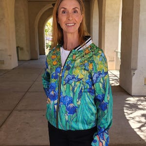 Puede incluir: Una vibrante chaqueta bomber con un diseño floral de iris azules, follaje verde y acentos amarillos. La chaqueta tiene una cremallera negra y un cuello a rayas blanco y negro. La chaqueta se usa con una camisa blanca y pantalones negros.