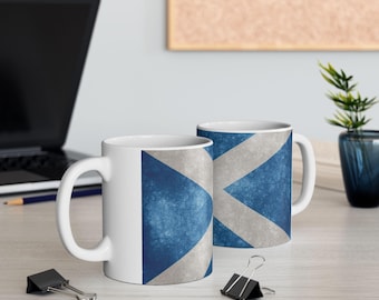 May include: Two white ceramic mugs with a blue and gray Scottish flag design. The flag features a white X-shaped cross on a blue background. The mugs are on a light-colored surface.