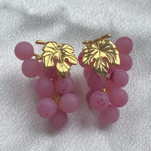 Könnte beinhalten: Ein Paar ohrringförmige Trauben. Jeder Ohrring hat eine Ansammlung von rosa, runden Perlen, die Trauben ähneln, mit einem goldfarbenen Blattakzent oben. Die Ohrringe sind vor einem weißen, strukturierten Hintergrund platziert.