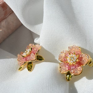 Könnte beinhalten: Zwei blütenförmige Ohrringe mit rosa und klaren, kristallartigen Blütenblättern, goldfarbenen Stielen und kleinen Perlenakzenten. Die Ohrringe liegen auf einem weißen Stoff, im Hintergrund ist ein rosa Samtkästchen zu sehen.
