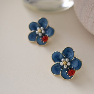 Op de afbeelding: Twee bloemvormige oorbellen met een marineblauwe emaille afwerking en gouden randen. Elke oorbel heeft een cluster van kleine witte parels in het midden en een kleine rode lieveheersbeestje. Op een witte achtergrond.