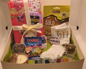 Puede incluir: Una caja de regalo llena de varios artículos, que incluyen un libro envuelto, una mascarilla facial, calcetines con flores amarillas, una vela, caramelos y pegatinas. Las pegatinas dicen "Mafia Romance Reader" y "Dark Romance Reader".