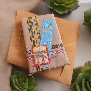 Puede incluir: Un paquete envuelto en papel marrón con cinta amarilla y negra de advertencia, cinta de cuadros rojos y blancos y un marcapáginas azul y blanco con el texto "Bee Happy!" y una ilustración de abeja. El paquete está sobre una superficie de papel marrón.