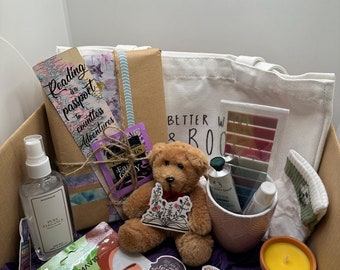 May include: A brown cardboard box filled with various items, including a tote bag with the text "Better With Books", a teddy bear holding a book, a face mask, a candle, and a book-themed bookmark.