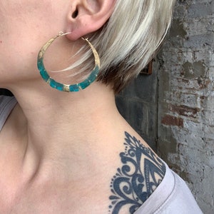 May include: Large gold hoop earrings with a teal blue enamel design. The earrings are worn by a person with short blonde hair.