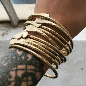 May include: A stack of gold-toned metal bangles with a hammered texture. The bangles are stacked on a person's wrist.